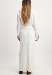 Pantalons larges à rayures blanches, avec une taille haute et des passants de ceinture. Une chemise à manches longues avec des détails aux poignets est également visible.