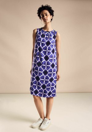 Robe sans manches bleue avec un motif géométrique en violet et noir. Tissu lisse avec un col froncé. Associée à des baskets blanches.