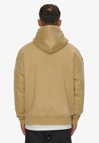 Beige hooded sweatshirt with a relaxed fit, featuring a large hood, long sleeves, and ribbed cuffs. Smooth texture and minimal design.