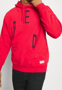 Sudadera roja con estampado de letras negras. Presenta un bolsillo tipo canguro, capucha ajustable y puños acanalados. Fabricada en una suave mezcla de algodón.