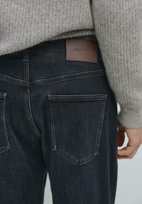 Jeans in denim scuro con etichetta in pelle recante la scritta "Massimo Dutti". Tasche posteriori visibili e dettagli di cucitura sottili. Maglione in lana parzialmente coprente.