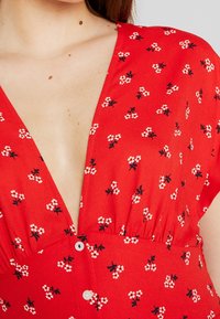 Vestido floral rojo con escote en V profundo, que presenta patrones de flores blancas y negras y detalles con pliegues en la cintura con un cierre de botón.