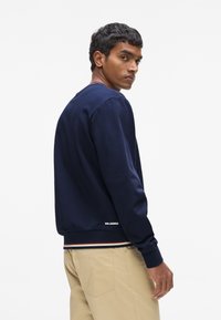 Sweat-shirt bleu marine avec poignets et ourlet côtelés, présentant des accents rayés en bas. Logo affiché au dos, coupe décontractée et texture lisse.