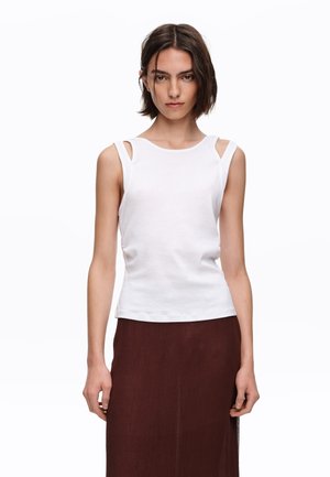 Joven mujer con cabello corto y oscuro lleva una blusa sin mangas blanca con recortes en los hombros y una falda larga marrón oscuro, de pie frente a un fondo blanco.