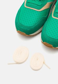 Vihreät tennarit, joissa on rei'itetyt varpaat ja beigeitä yksityiskohtia, asetettu kahden rullatun valkoisen kengännauhan päälle valkoista taustaa vasten.