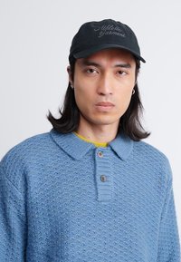 Svijetloplavi strukturirani džemper s ovratnikom i tri dugmeta; crna kapa s vezom teksta; osoba ima dugu kosu i naušnice.