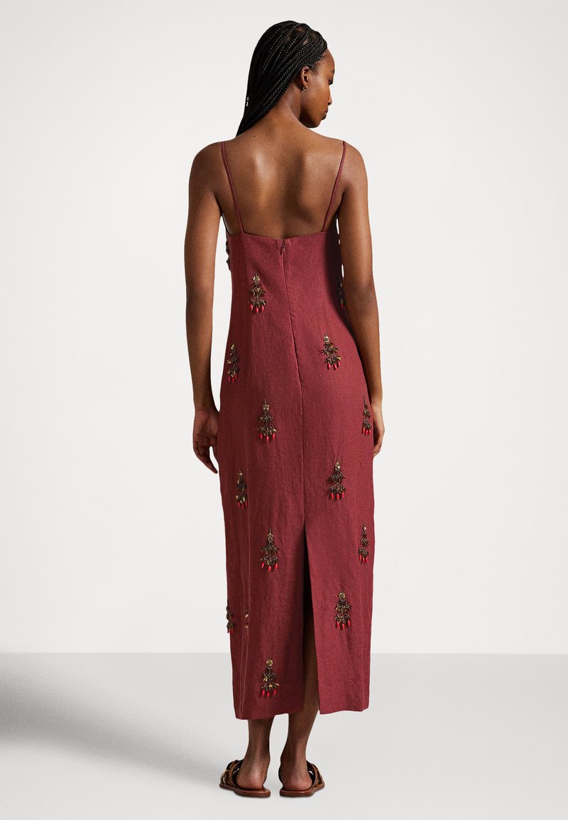 Frau mit geflochtenem Haar, die ein ärmelloses, bordeauxfarbenes Midikleid mit bestickten Blumenmustern und offenem Rücken trägt, vor einem schlichten Hintergrund stehend.