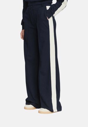 Pantaloni blu navy a gamba larga con strisce laterali bianche in rilievo, indossati con scarpe da ginnastica bianche; mano nella tasca visibile da un lato.