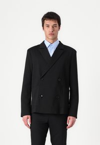 Schwarze zweireihige Jacke mit einem strukturierten Muster, zwei vorderen Taschen und schwarzen Knöpfen, getragen über einem hellblauen Hemd.
