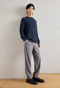 Pulover tricotat bleumarin, pantaloni gri pliați și pantofi negri. Puloverul are guler rotund; pantalonii au buzunare laterale și un croi lejer.