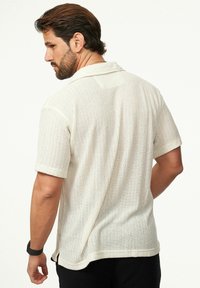 Polo mit kurzem Arm aus leichtem cremefarbenem Strickstoff mit strukturiertem Muster; verfügt über einen Kragen und Seitenschlitze am Saum für eine bessere Bewegungsfreiheit.