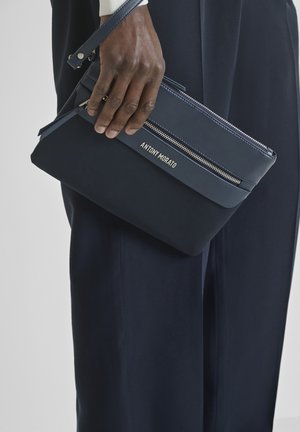 Mano sosteniendo un bolso pouch con cremallera Antony Morato azul marino sobre un fondo de pantalones oscuros a juego.