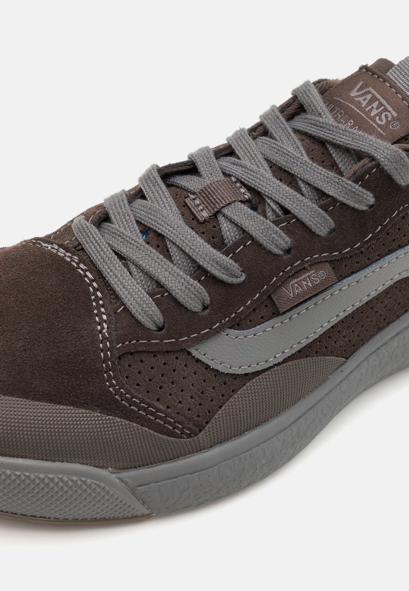 Zapatillas de ante marrón con detalles grises, perforaciones, cordones grises y suela de goma. Cuenta con una etiqueta de marca en el lado.