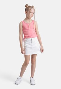 Νεαρή κοπέλα με διπλούς πλεξούδες σε σχήμα κότσου, που φορά ροζ μπλούζα χωρίς μανίκια, λευκή φούστα και λευκά αθλητικά παπούτσια, μπροστά σε απλό λευκό φόντο.