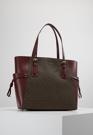 Sac cabas Michael Kors marron et vert foncé avec doubles anses, ferrures dorées et détails de lanière sur les côtés, posé sur une surface blanche.