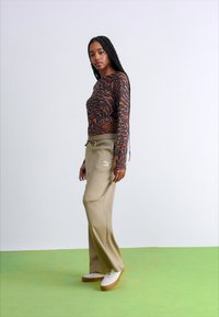 Top lung cu mâneci lungi de culoare maro, cu un model spiralat portocalie, asortat cu pantaloni de trening bej deschis, cu șnur și logo. Modelul stă pe un podea verde.