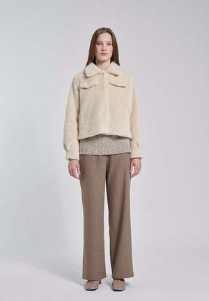 Giovane donna in piedi che indossa una giacca crema pelosa, un maglione beige, pantaloni marroni a gamba larga e scarpe beige su uno sfondo grigio chiaro.