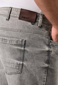 Jeans in denim grigio con un'etichetta in pelle "MEN+" sul retro. Presentano un design standard delle tasche e una leggera sfumatura nella trama.