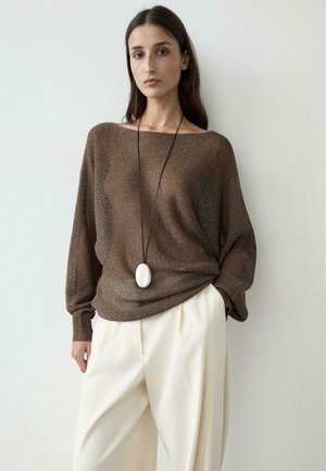 Donna che indossa un maglione marrone largo a maglia, pantaloni color crema a gamba larga e una lunga collana nera con un grande pendente ovale bianco.