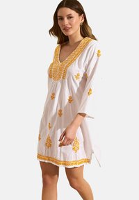 Robe en coton blanc avec un col en V, ornée de broderies florales jaunes et de garnitures décoratives. Manches longues et légèrement au-dessus du genou.