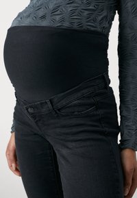 Zwarte rekbare zwangerschapsjeans met een fitted tailleband en knoopsluiting, gecombineerd met een gestructureerde grijze blouse met lange mouwen die een patroonontwerp toont.