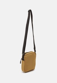 Bruine stoffen crossbodytas met een rechthoekige vorm, ritsluiting en een zwarte verstelbare schouderriem. Soepele textuur met subtiele stiksels.