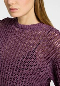Woman wearing a purple, loosely knit sweater over a black top, showing texture and open knit pattern on the fabric.