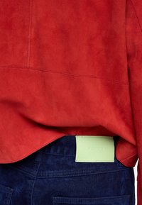 Czerwona zamszowa kurtka częściowo przykrywająca ciemnoniebieskie dżinsy z jasnozieloną prostokątną metką przyszytą nad tylną kieszenią.