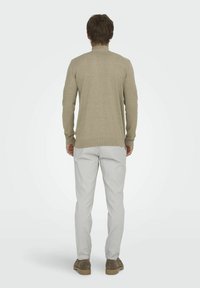 Helle beige Strickjacke mit einem gerippten Kragen und Bündchen, kombiniert mit hellgrauen Hosen und braunen Schuhen, von hinten betrachtet.