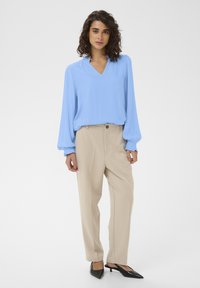 Blouse bleu clair avec manches longues froncées et col en V ; associée à un pantalon beige ; talons noirs à bouts pointus. Design simple et moderne.