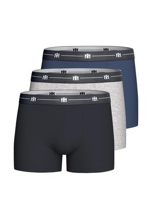 Uomo senza maglietta con pelle chiara e capelli castani che indossa boxer grigi, sistema la cintura, in piedi davanti a uno sfondo chiaro e uniforme.