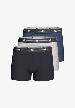 Man zonder shirt met een lichte huid en bruin haar, draagt grijze boxershorts, past de tailleband aan, staand tegen een effen lichte achtergrond.
