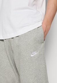 Acercamiento de una persona que lleva pantalones de chándal gris claro de Nike con la mano en el bolsillo y una camiseta blanca.
