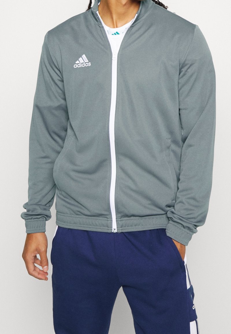 Grå zip-up jakke med høj krave, hvid lynlås og Adidas-logo på venstre bryst. Blødt, letvægtsstof.