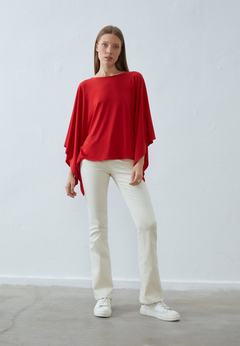 Top rosso, leggero, a taglio morbido con maniche ampie; abbinato a pantaloni aderenti color crema e sneakers bianche. Texture liscia, design minimale.