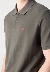 Vyriška polo marškiniai tamsiai žaliame medvilnės audinyje, su klasikinio tipo apykakle, dviem sagomis ir kontrastinga rožine logo ant krūtinės.