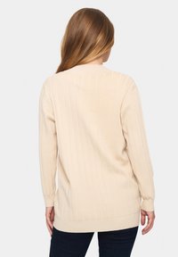 Bežinė raukšlėta megztinė su ilgomis rankovėmis, apvaliu iškirpimu ir laisvu siluetu. Audinys atrodo minkštas ir tekstūruotas.