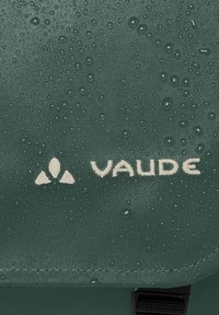 Tessuto impermeabile verde con gocce, presenta il logo bianco "VAUDE" e un emblema triangolare. Texture liscia e dettagli di bordo cucito visibili.