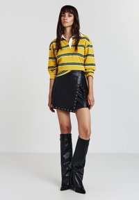 Frau trägt ein gelb gestreiftes Langarmshirt, einen schwarzen Leder-Minirock mit Ösen-Details und kniehohe schwarze Lederstiefel vor einem schlichten Hintergrund.