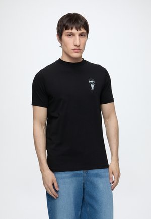 Jeune homme portant un t-shirt noir avec un petit motif d'astronaute sur la poitrine et un jean bleu, debout devant un fond blanc uni.