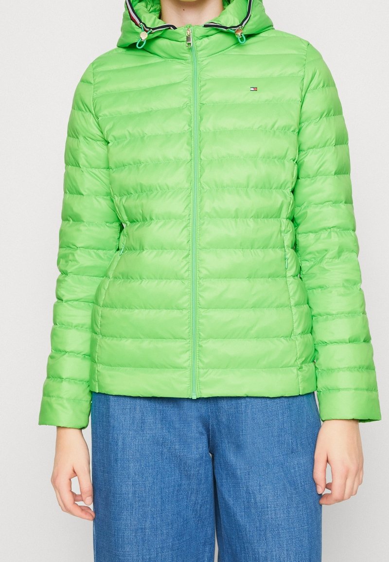 Groene gevoerde jas met capuchon, doorgestikte textuur, ritssluiting aan de voorkant en klein logodetail. Gecombineerd met blauwe broek.