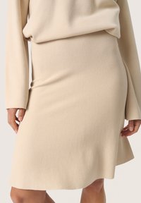 Strik lavet af beige garn med en glat tekstur, tætsiddende talje og flagrende kant. Designet med lange ærmer og en afslappet pasform ovenfor.