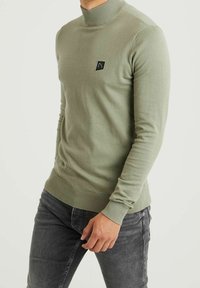 Pull à col roulé vert olive avec poignets et ourlet côtelés, comportant un petit patch logo noir sur la poitrine, porté avec un jean foncé.