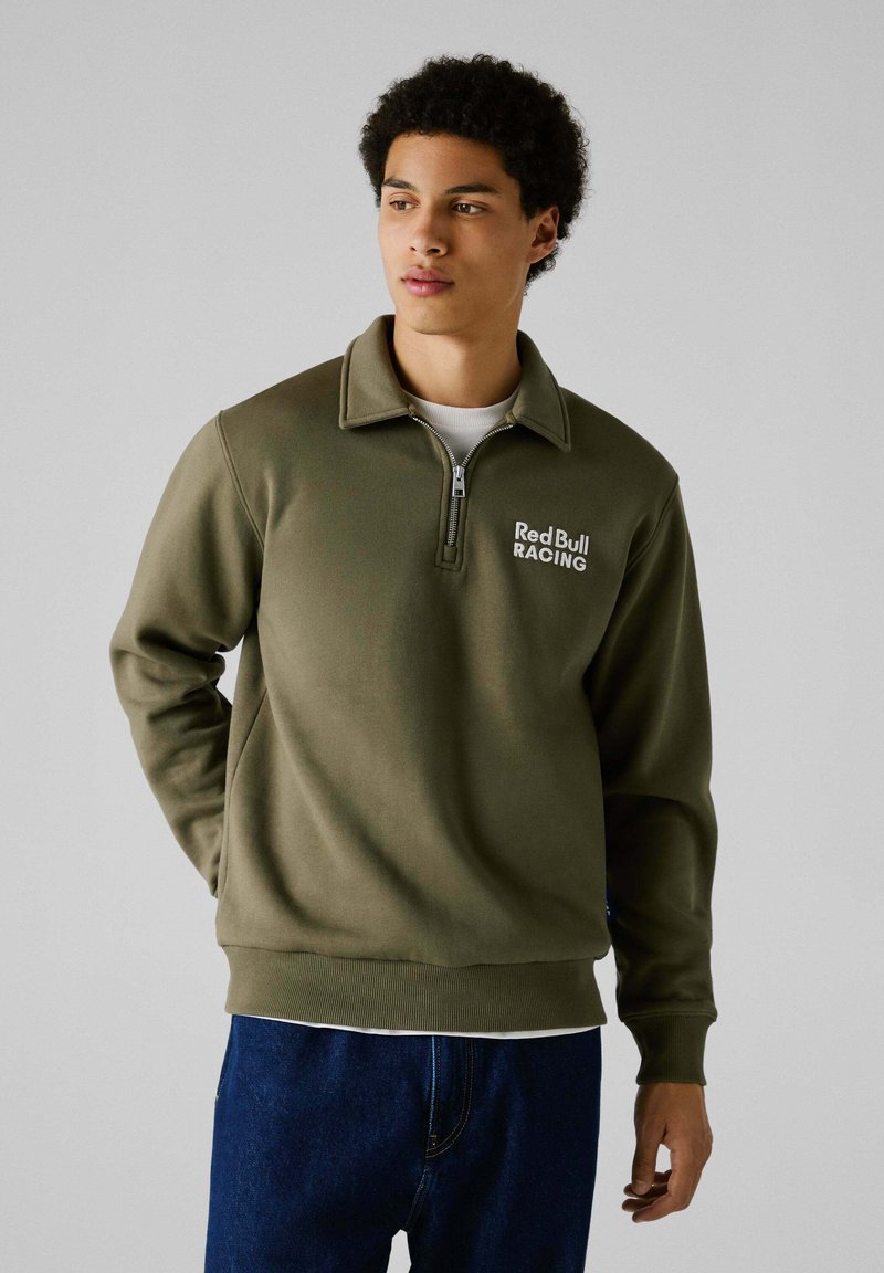 Sudadera de color verde oliva con cuello con cremallera, que presenta el logo "Red Bull RACING", hecha de una tela suave, combinada con jeans de mezclilla azul.