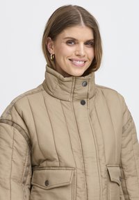 Beige quiltet jakke med høj krave, der har to frontlommer og sorte trykknapper, lavet af et tekstureret materiale.