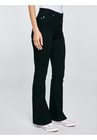 Sieviete, kura valkā melnas zvana formācijas džinsas un baltas sporta kurpes, stāv sānis pret vienkāršu gaišu fonu.