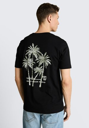 Man draagt een zwart T-shirt met een afbeelding van vijf palmbomen en tekst op de achterkant, gecombineerd met lichtblauwe spijkerbroek, staand met het gezicht naar rechts.