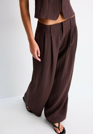 Femme portant un pantalon large marron foncé et un gilet court assorti, debout avec une main détendue le long du corps et des sandales noires aux pieds.