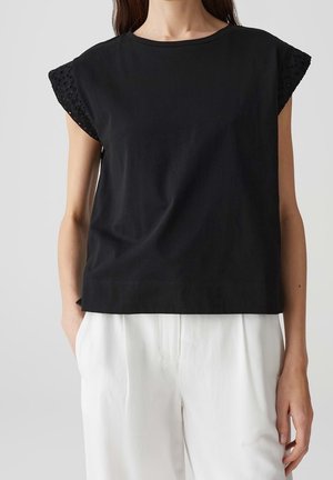 Femme portant un haut noir sans manches avec un détail en dentelle œillet sur les manches et un pantalon blanc ample sur un fond uni.