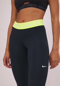 Leggings de sport noirs avec une large ceinture néon jaune portant l'étiquette "NIKE PRO". Le tissu semble lisse et extensible, conçu pour les vêtements de sport.